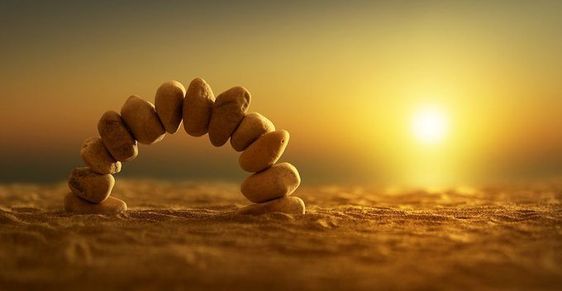 A collection of small stones on a beach balanced perfectly upright in an arch with the sunsetting in the background. Photographed by Anna Ovatta