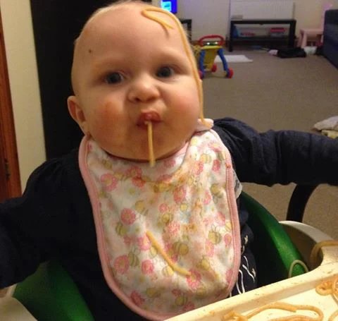 A baby sitting in a high chair eating spaghetti some is on his head and some sucked up on in his house