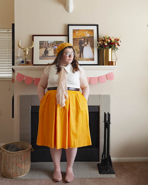 An outfit consisting of a yellow beret, oversized white ruffled peter pan collar with a pastel pink lace scarf tied under under a white short sleeve lace blouse tucked in to a yellow midi skirt and dusty pink pointed toe mules.