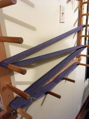 A square wooden warping board with long pegs on a white wall, with four spans of grey yarn and a narrow band of teal looped across it, tied with yellow cord.