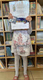 Na zdjęciu nagrodzona dziewczynka trzymająca w dłoniach dyplom oraz nagrodę. Dziewczynka stoi na tle regału z książkami, ma zasłoniętą twarz.