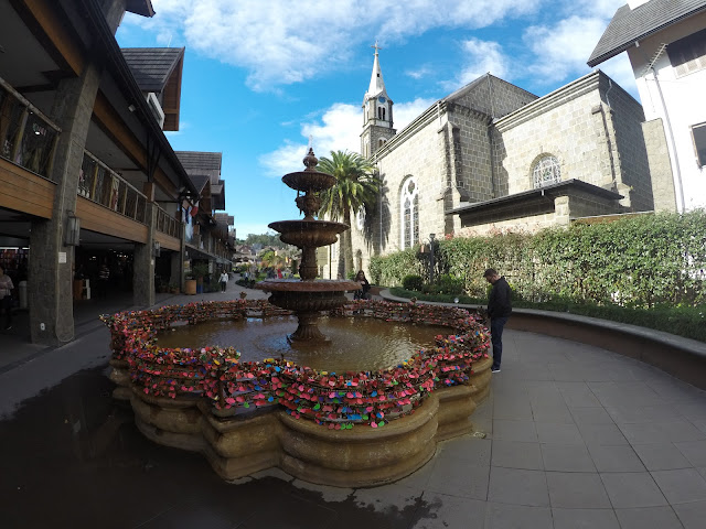 Fonte com um pequeno chafariz, cercada por uma grade baixa, onde os turistas apaixonados costumam encher de cadeados com etiquetas coloridas, onde constam seus nomes, ou mensagens de amor. Ao fundo à direita, a igreja de pedra, de Gramado.