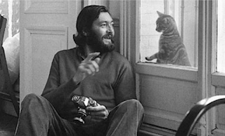 Julio Cortázar con cámara fotográfica en mano, sentado en el piso, dialogando con un gato