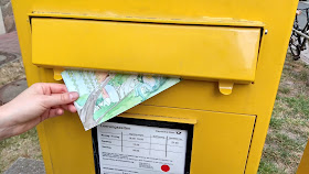 Das Foto zeigt die Hand einer Frau, die eine für Kinder illustrierte Postkarte in einen großen gelben Briefkasten der Deutschen Post einwirft.