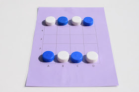 na zdjęciu widać planszę do gry Tac Tickle. Plansza jest w kolorze fioletowym i ma dwadzieścia pól oznaczonych w poziomie literami a w pionie cyframi. Na planszy leżą pionki, cztery w kolorze białym i cztery w kolorze niebieskim, naprzemiennie po obu stronach planszy. To ustawienie wyjściowe.