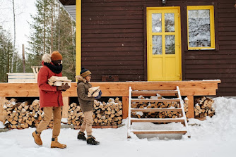 A man and boy carrying firewood. They are in front of a house with a yellow door that has a large front porch with firewood underneath it.