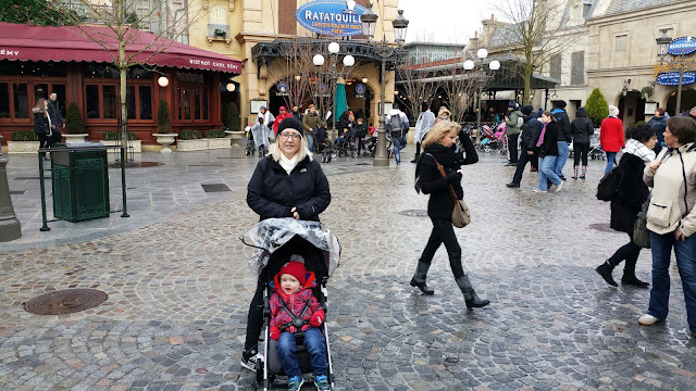 Little boy in a pushchair with his Mum standing behind it and the Ratatouille area in the background