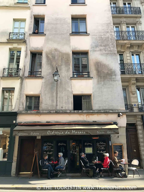 Five people sitting at tables in front of a Crêperie in the Marais on a sunny afternoon.