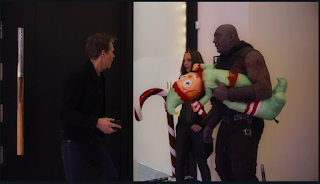 Kevin Bacon standing at his door, Drax and Mantis standing in his entry way. Drax is holding an inflatable elf and Mantis has a giant candy cane.