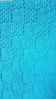 A photo of part of a knitted blanket. it has a square pattern like a chess board. some squares and the border are lines of knitting and some are filled with stitches in a curved pattern.
