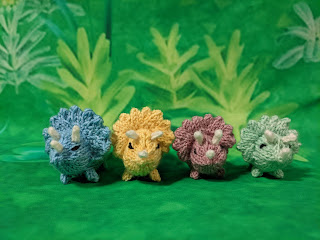 Green plant and grass background with four, handknit, small triceratops, pastel blue, pastel yellow, pastel muave, and pastel mint