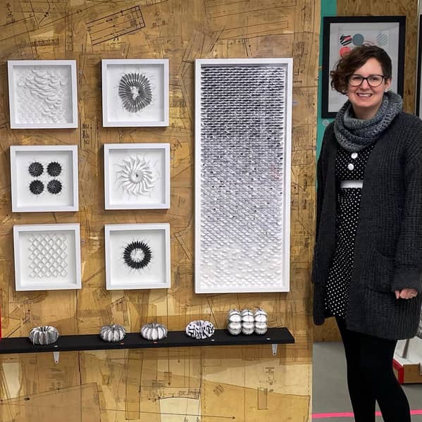 smiling woman wearing black and white winter clothing stands next to wall of black and white stitched and framed paper art with sculptural paper objects on shelf below