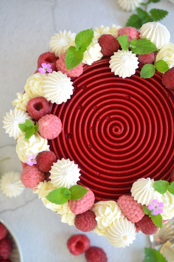 Himbeer-Dirndlmousse-Torte mit Brennnessel-Biskuit von oben fotografiert – zu sehen ist die tiefrote Himbeerspirale und rundherum Schlagoberstupfen, frische Himbeeren und Himbeeren aus Dirndlmousse, Baiser, Blätter und Blüten.