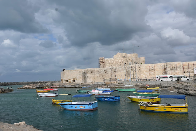 A panoramic view of the new Qaitbay Promenade in Alexandria, Egypt, with visitors walking along the 550-meter sea walkway beside the historic Qaitbay Citadel during sunset.