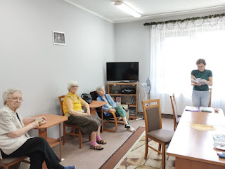 Tło: pokój z dużym oknem. Zdjęcie przedstawia trzy panie seniorki i panią bibliotekarkę. Panie seniorki siedzą na krzesłach i fotelu, pani bibliotekarka stoi przy prostokątnym stoliku. Na wprost stoi wyłączony telewizor na okrągłym stoliku.