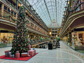 A photo of the inside of a shopping mall decorated for Christmas, on the top floor, taken from the right corner of a length of shops. A Christmas tree with presents around the bottom of it is on the left hand side, just in front of a long opening in the floor, where shoppers are coming up on an escalator.