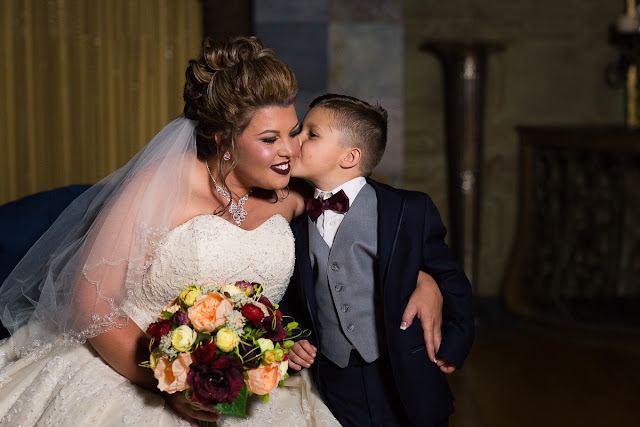  Cassie and Wyatt tied the knot at The Palace Event Center, in Yukon, OK, on July 15, 2017.   The venue’s magnificent wood entry doorways, outside rock façade, and adjacent country landscape provided the perfect backdrop to photograph the first look and pictures of the jovial wedding party.  The venue was spacious, which allowed us to sneak in a few pre-dressed shots of the girls in their “Wedding Tribe” t-shirts.     Cassie’s elegant, form-fitting, strapless, satin wedding dress overlaid with lace and sequins looked like it was made especially for her.  Wyatt was quite the Knight in Shining Armor dressed in his navy suit, grey vest, and burgundy tie.  The four beautiful bridesmaids wore matching full-length, flowing, burgundy gowns, slit at the knee.  Cassie’s gorgeous bouquet was composed of baby’s breath, peonies and ranunculus in rich hues of vibrant burgundy, red, pink, burnt orange, and yellow gold.  To add to the excitement of the day, their son had a special assignment—carrying the sign down the aisle that stated, “Daddy, Here Comes Our Girl.”     The venue’s grand ballroom, with it’s beautiful chandeliers and romantic lighting, was the perfect backdrop for the couple’s reception.  Candlelight, flowers, and twinkle lights adorned the nuptial stage, and after the vow exchange, Cassie and Wyatt participated in their first dance.  Guests were invited to enjoy the buffet, the cutting of the cake, and the toasts by the best man and bridesmaid.  Chairs were quickly removed from the dance floor, and while almost everyone spent the rest of the afternoon relaxing, dancing and socializing, there was a particularly fun-loving group of guests who decorated the bride and groom’s Jeep with sayings, crepe paper, and a car filled-to-the-brim with balloons!        Cassie stated that the best part of the wedding was being able to see so many family and friends at the reception having fun and enjoying themselves.  She said, “If we had it to do over, we wouldn’t do anything differently…it was perfect for both of us!”     It was a pleasure to capture the special moments of this memorable wedding day for Cassie and Wyatt, and we wish them a lifetime of love and happiness. 