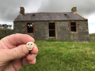 A photo of a small ceramic skull (Skulferatu 88) being held up with the ruined farmhouse of Upper Bardennoch Farm in the background.  Photo by Kevin Nosferatu for the Skulferatu Project.