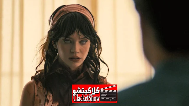 الصورة تبدو وكأنها مشهد من فيلم غامض أو رعب. تظهر امرأة ذات نظرة زائغة وعينين شاحبتين وكأنها مسحورة أو تحت تأثير قوة خارقة. ترتدي وشاح رأس وملابس تبدو ذات طابع قديم أو مستوحى من السبعينيات. في المقدمة، هناك شخص آخر يظهر من الخلف، مما يضيف إلى جو التوتر في المشهد. الإضاءة الدافئة والخلفية الضبابية تعطي إحساسًا بالغموض. في أسفل الصورة، يوجد شعار "كلاكيتشو ClacketShow"، مما يشير إلى أنها جزء من ملخص فيلم.