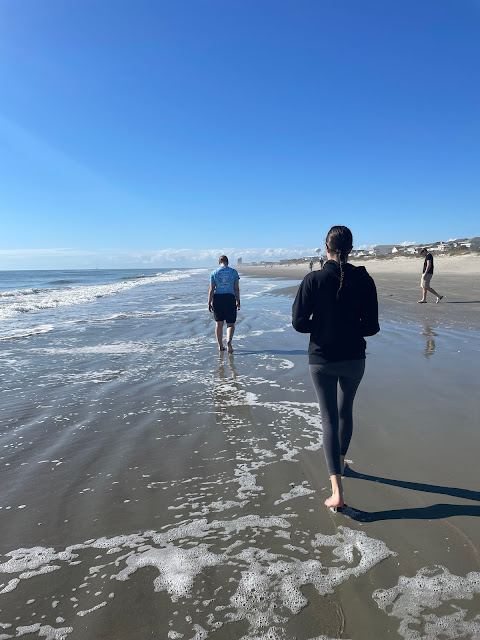 You and Elizabeth taken from behind walking down the beach on our long walk. The water is on your left. The waves are gentle and the water is slowly coming up to our feet. On the left is the housing for the town. The sky is blue and the sun is shining on the left hand side of our bodies.