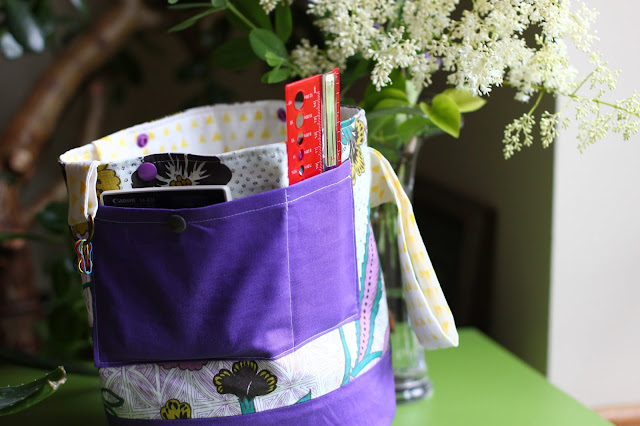 Reverse side of purple floral project bag features a dual purple pocket that's holding a calculator and a ruler in the image. Top small loop has a D-rinng holding removable stitch markers. 