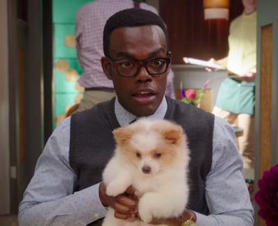 Chidi is sitting in a cafe, with some people in the background, and he's holding a small, very fluffy white and tan puppy.