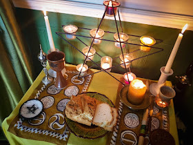 Green painted paneled wall with white dado-rail. Leaf-green curtains of synthetic satin. The altar-cloth is yellow, and on it are two gold place-mats with La Tène style designs, a pentagram candle-holder, an earthenware chalice, a pentacle dish with salt, a gold candle in an earthenware dish, two white soy taper candles in off-white candle-sticks, and a loaf of home-made bread