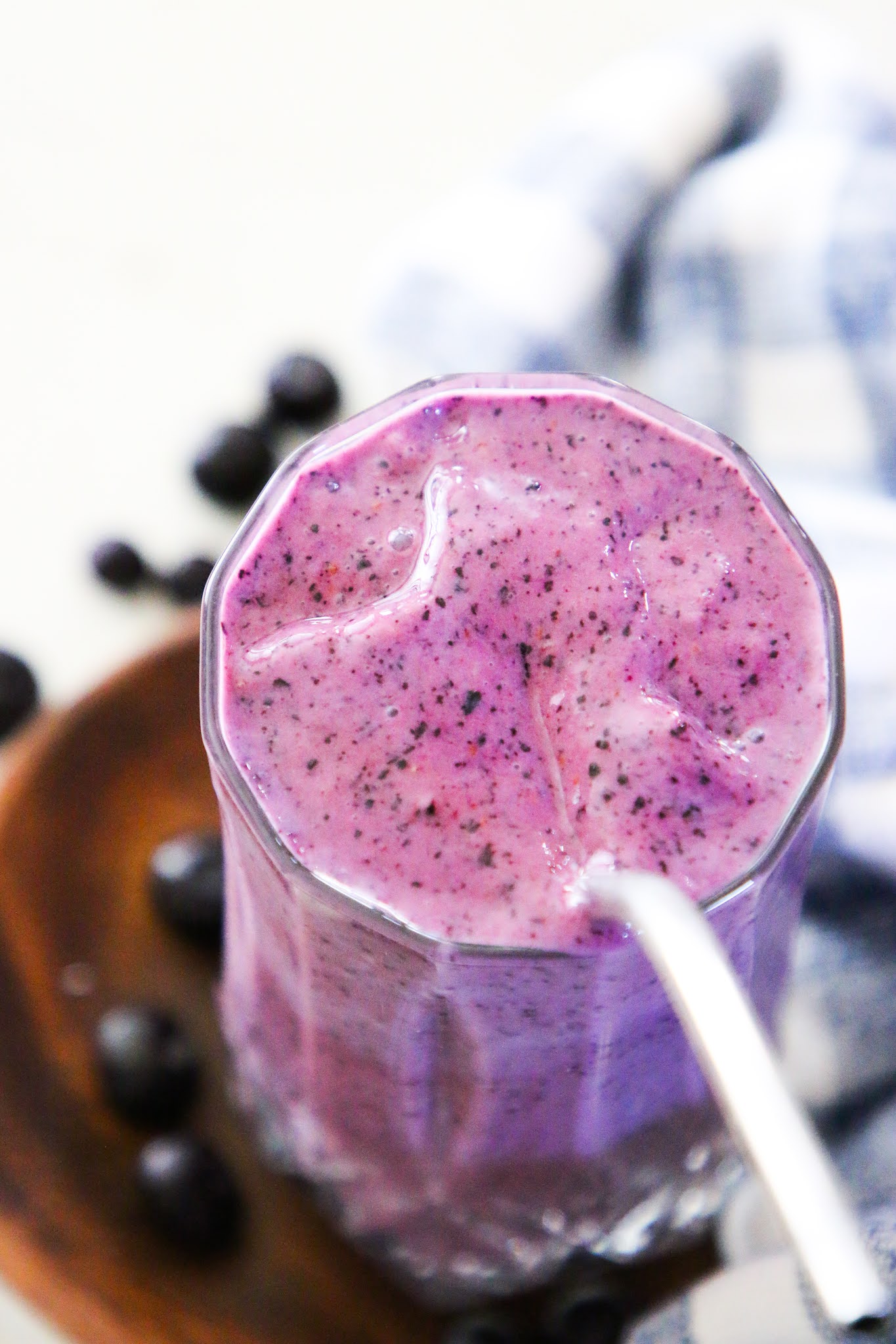 Purple blueberry smoothie in a tall glass with blueberries on top. There is a wood coaster underneath the smoothie sitting on top of a white marble table. The glass is wrapped in a blue dishcloth.