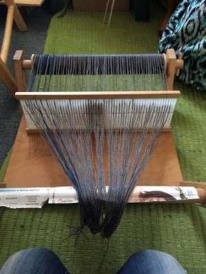 A small wooden rigid heddle loom, with the grey-and-teal warp wound onto the back beam and the heddle sleyed. the ends of the warp threads are draped over the front beam, and the loom is sitting on a green rug.