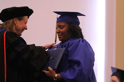 Two women in graduation gowns and hats. One presenting an online Bachelor's degree to another woman.