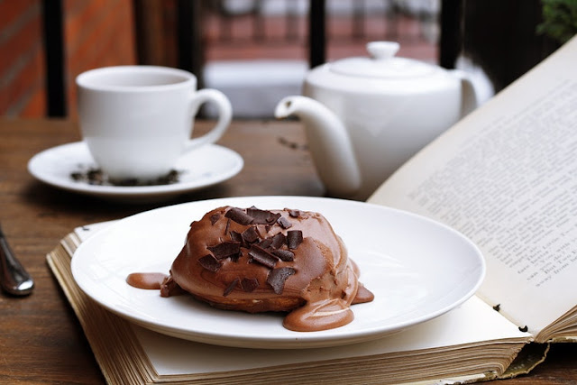 Chocolate cake on a plate along with a book and teacup and saucer