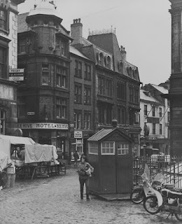 A black and white photograph of a wet cobbled marketplace. A police call box is at the centre of the image with a person walking by carrying something. There is a row of shops including an old-fashioned Hotel in the background, with the rear of a covered market stall in the middle ground.
