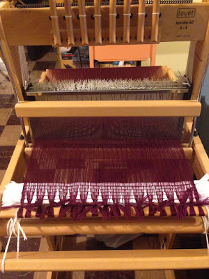 An eight-shaft table loom, warped across the entire width with maroon, and with several rows of toilet paper packed into the beginning to spread the warp threads.