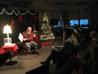 A white bearded man in flannel reads a picture book to an audience of adults and children with a lit Christmas tree to his right and a lamp on a table to his left.