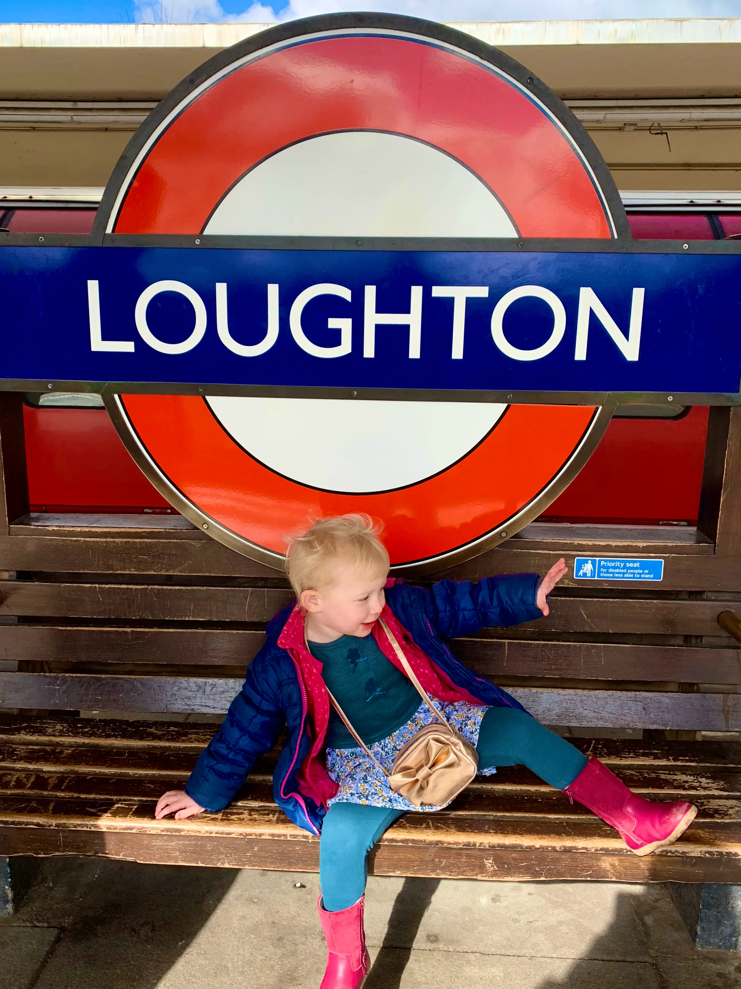 A child sitting on a bench next to Loughton underground station sign