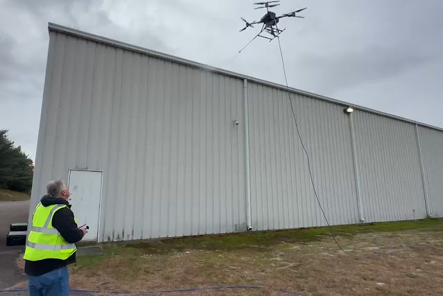 A transportation worker navigating a midsize black drone hovering in the air next to a commercial storage building. A long spray nozzle is attached to the front of the drone