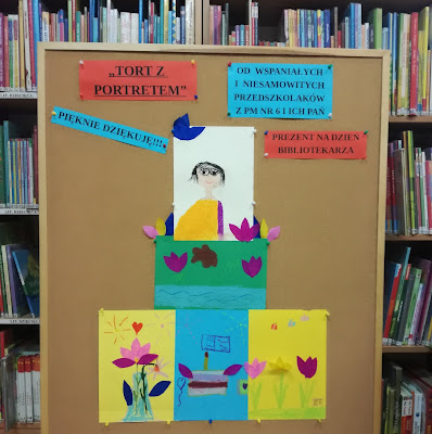 Prezent na dzień bibliotekarza praca plastyczna Tort z portretem od przedszkolaków z PM nr 6 na korkowej tablicy na tle regałów z książkami