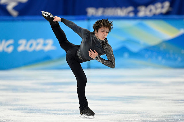 Uno Shoma patina com apenas um patim, segurando o pé esquerdo atrás do seu corpo. Ele veste uma calça preta e blusa cinza