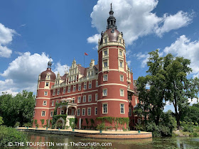 Water lilies on a pond in front of a red-ish coloured renaissance castle with an overgrown balcony under a blue sky with fluffy clouds