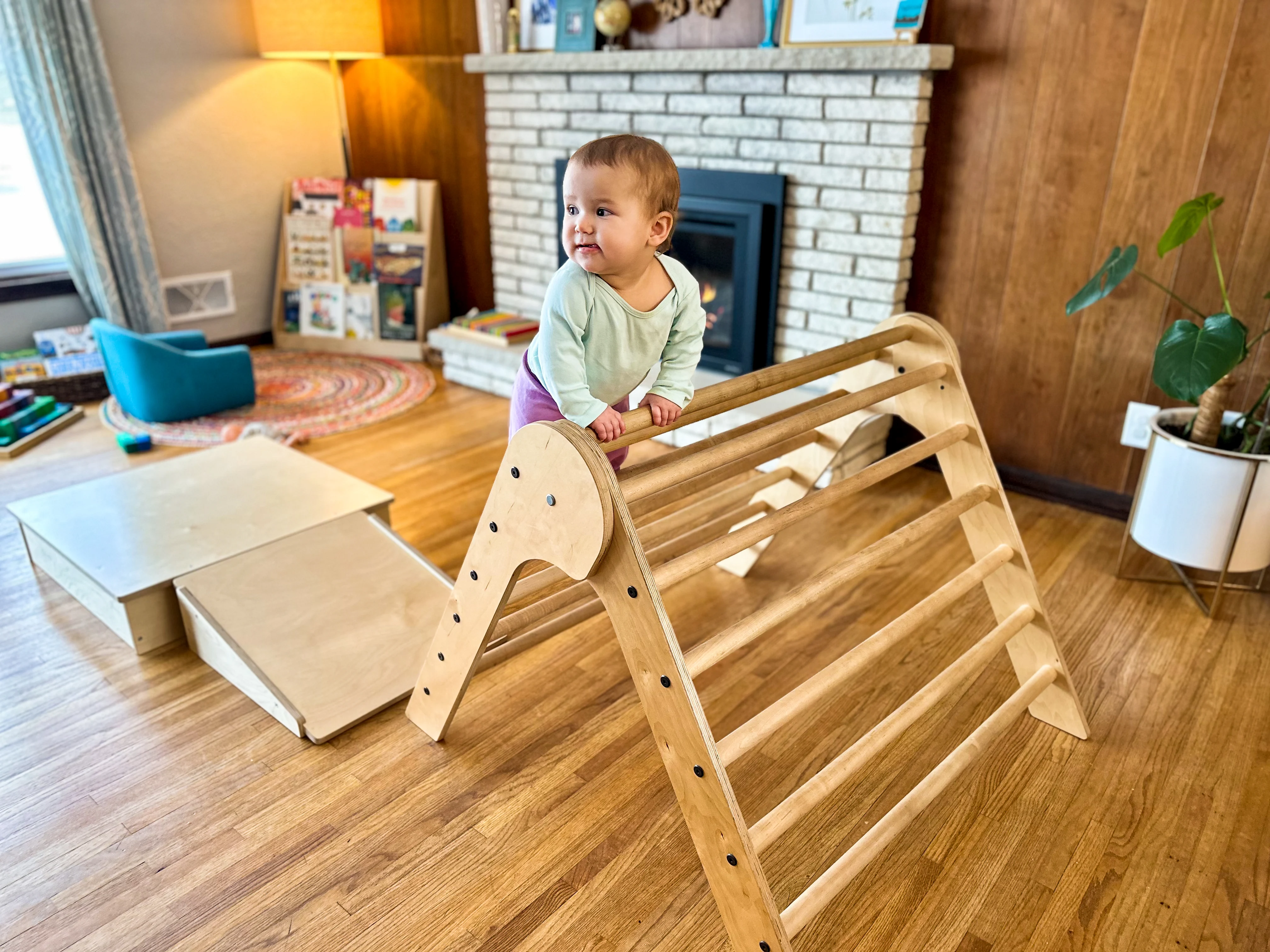 11-month-old Montessori baby climbs half way up a Pikler Triangle in her Montessori home. A Pikler ramp and platform can be seen in the background with a child friendly reading area.