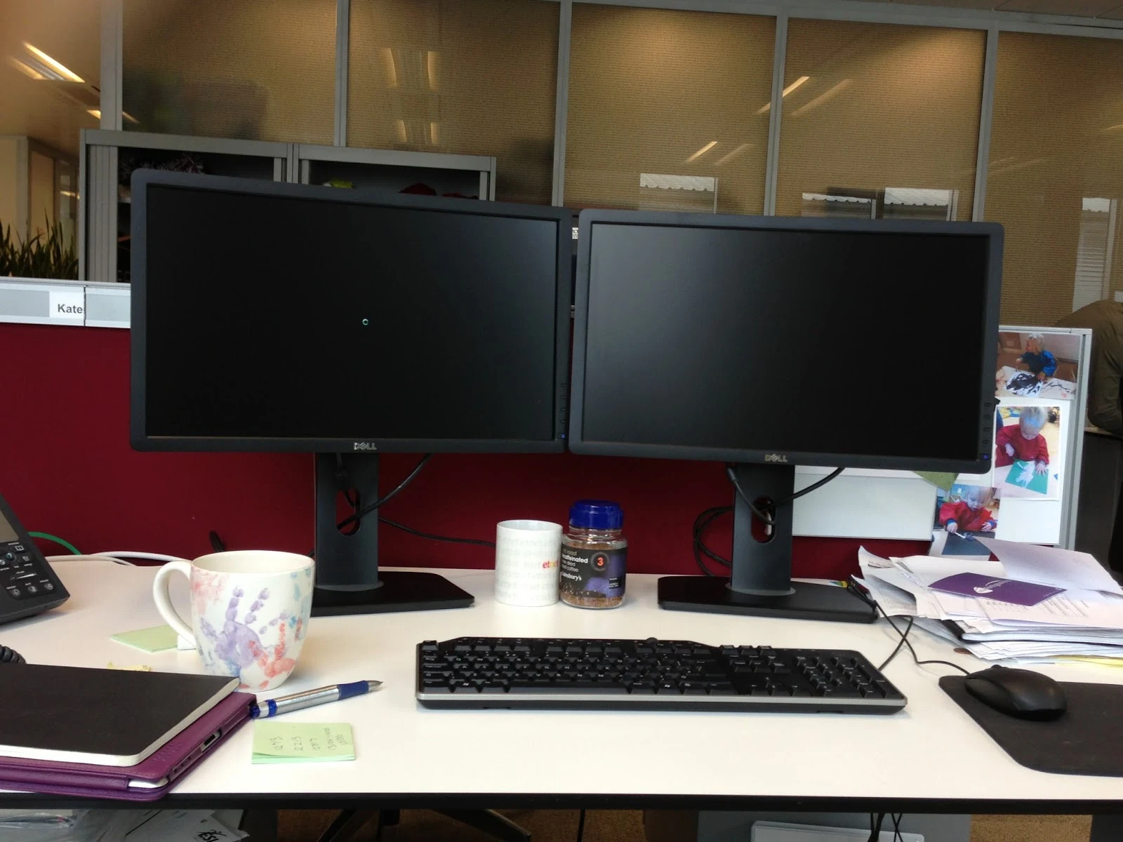 A desk in an open plan office with two monitors, phone and other desk stuff