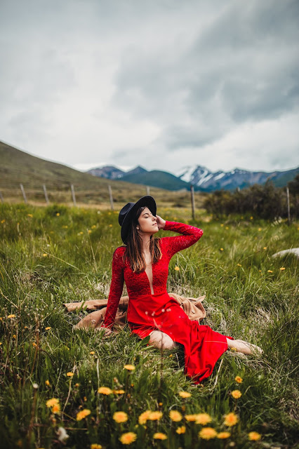 Uma mulher branca com um vestido longo vermelho e um chapéu preto. no fundo uma paisagem com grama verde. 