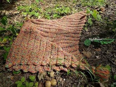 Na leśnej polanie leży melanżowa (odcienie fioletu, zieleni i pomarańczy), szydełkowa, trójkątna chusta. Jeden z jej rogów został zagięty, aby pokazać obie strony chusty na jednym zdjęciu.