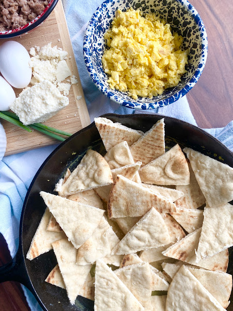Overhead shot of cast iron skillet with pita bread triangles & wooden cutting board with breakfast nacho ingredients.