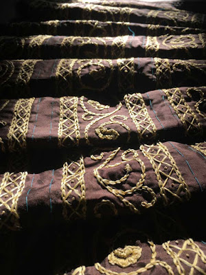 Three long bands of gold scrolling oak leaf embroidery on deep brown silk, with narrow chain-stitch and diamond borders on each band of oak leaves, and turquoise basting stitches marking the edges of each strip. The base fabric is softly pleated across the bands, horizontal to the viewer, making dramatic highlights and shadows on the folds from the strong sunlight coming in from above.