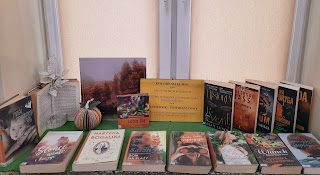 Na fotografii znajduje się czternaście książek z pomarańczowym kolorem na okładce, informacja o akcji umieszczona w ramce, grafika na kartce przedstawiająca jesienny las oraz ozdoba zrobiona ze starej książki przedstawiająca drzewo i dynię. Książki są ułożone na zielonej serwecie na parapecie okna.