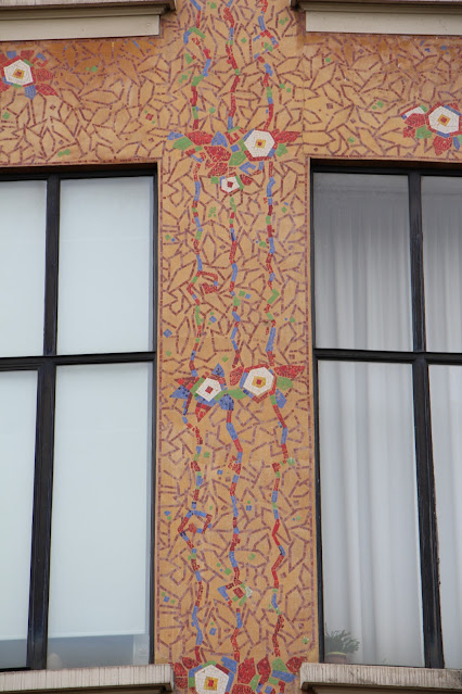 Photo. Detail of two window frames and the brickwork between and around the windows. The detailing is multicolored geometric vines and flowers.