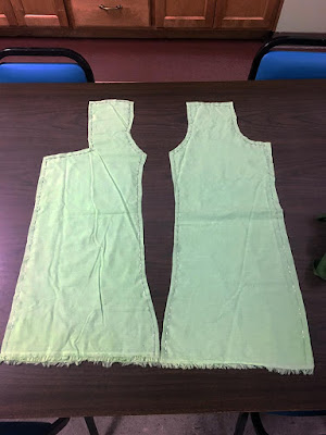 Two pale-green shirt pattern pieces, laid flat on a brown break room table and pinned densely around the outer edges.