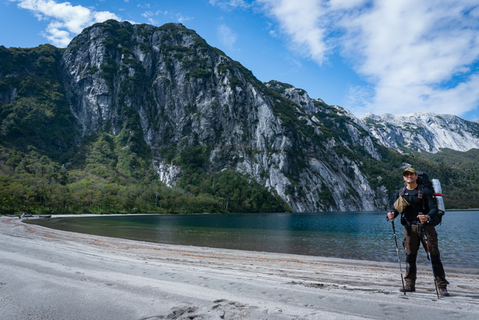 A man is standing on the beach in front of the water with hiking sticks and a backpack.