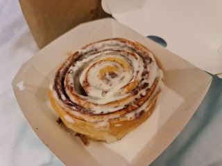 A photo of a circular light brown cinnamon roll with brown sugar in the middle coated in a white thick icing on a bright background